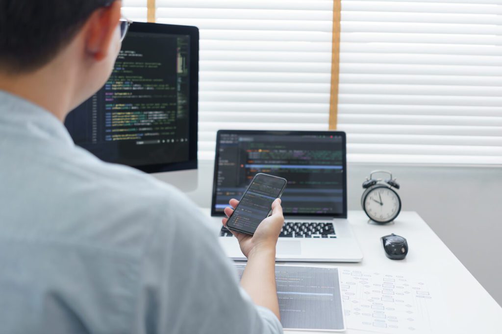 Man seated in front of desktop, laptop and clock, while holding a mobile device in hand